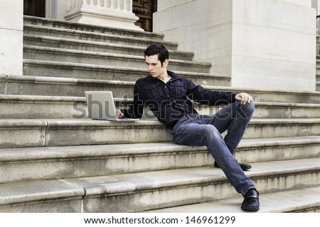 A young handsome guy is sitting on steps and studying on a computer. / Study Outside