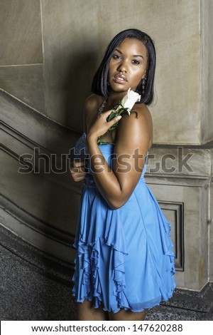 Dressing in a blue strapless skirt dress and holding a white rose, a black teenage girl is standing by the wall and tenderly looking at you, lost in thought. / Black GIrl and White Rose