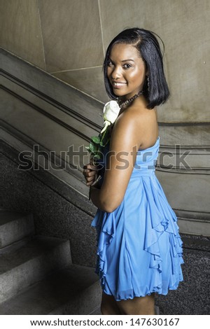 Dressing in a blue strapless skirt dress and holding a white rose, a black teenage girl is smiling and looking at you. / Black GIrl and White Rose