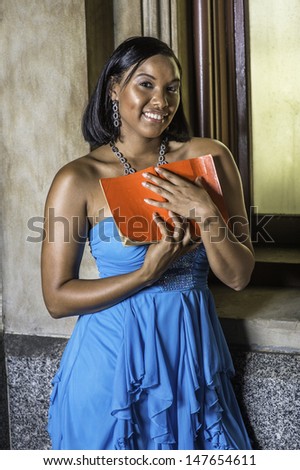 Holding a red book, smiling,  a pretty black girl is standing by the window, reading outside. / Reading By the Window