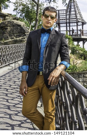 Dressing in a black blazer, a blue shirt, brown pants and a black bow tie,  a young businessman wearing sunglasses is standing in a bridge, relaxing. / Traveler