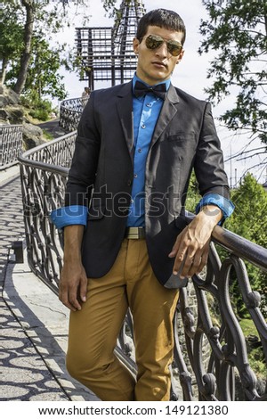 Dressing in a black blazer, a blue shirt, brown pants and a black bow tie,  a young businessman wearing sunglasses is standing on a bridge, relaxing. / Traveler
