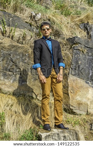 Dressing in a black blazer, a blue shirt, brown pants and a black bow tie,  a young businessman wearing sunglasses is standing on rocks and watching. / Watching