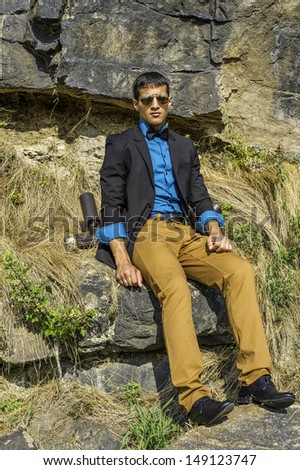 Dressing in a black blazer, a blue shirt, brown pants and a black bow tie,  a young businessman wearing sunglasses is sitting on rocks and relaxing. / Relaxing Outside