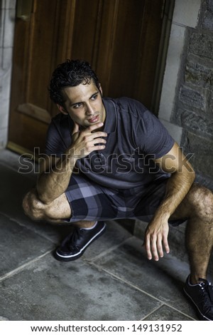 Dressing in a gray polo shirt, pattern shorts and black leather sneakers, one hand touching his chin,  a young strong handsome guy is squatting in the front of the door, relaxing and thinking.