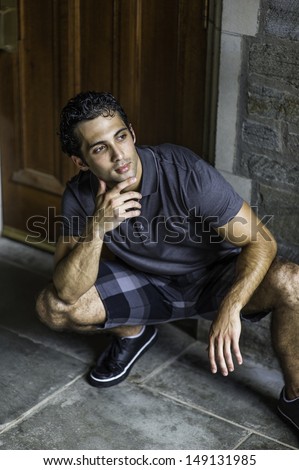 Dressing in a gray polo shirt, pattern shorts and black leather sneakers, one hand touching his chin,  a young strong handsome guy is squatting in the front of the door, relaxing and thinking.