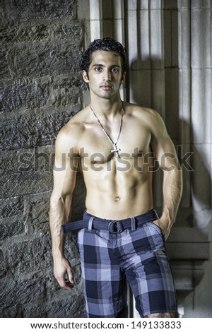 Wearing a cross necklace and half naked, a handsome, muscular guy is standing by old fashion rocky wall, confidently looking at you. / Portrait of  Fitness Guy