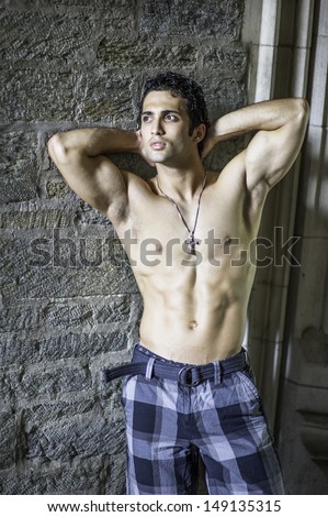 Wearing a cross necklace and half naked, a handsome, muscular guy is standing by old fashion rocky wall and relaxing. / Portrait of  Fitness Guy