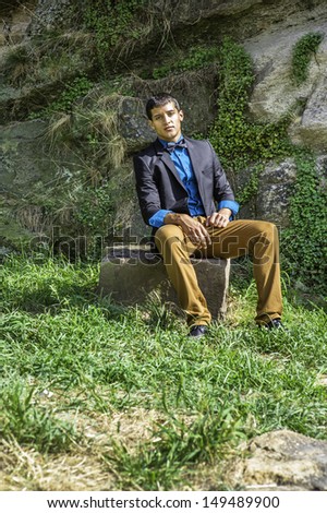 Dressing in a black blazer, a blue shirt, brown pants and a black bow tie,  a young businessman is sitting on rocks and taking a break. / Taking Break