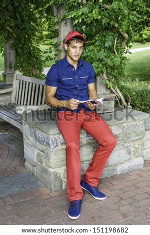 Dressing in a red baseball hat, a blue short sleeve shirt,  red pants and blue shoes, a handsome, attractive guy is reading a book outside. / Reading Outside
