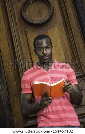 Dressing in a red, pink lines T shirt, wearing a bracelet, a young black guy is reading a red book outside. / Black Guy and Red Book