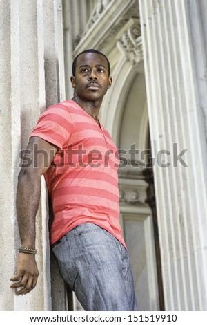 Dressing in red, pink lines T shirt,  gray pants, wearing a bracelet, a young black guy is standing by a pillar, confidently looking forward. / Portrait of Young Black Guy