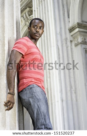 Dressing in red, pink lines T shirt,  gray pants, wearing a bracelet, a young black guy is standing by a pillar, confidently looking forward. / Portrait of Young Black Guy