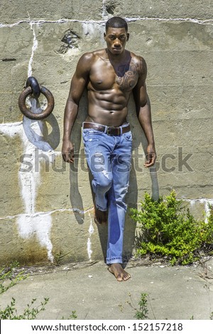 Under the sunshine, a masculine black guy, wearing blue jeans, barefoot and half naked, is standing by a wall, confidently looking forward. / Portrait of  Young Black Fitness Guy