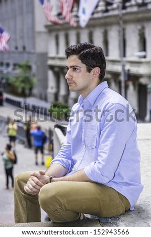 Clamping his hands, a young handsome guy is sitting outside on a stage into deeply thinking. There are American flags hanging on buildings in the background. /Thinking Outside