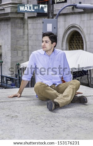 A young handsome guy is sitting on a stage in the corner of the street, relaxing and thinking. There is a Wall Street sign in the background. / Thinking and Relaxing outside