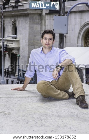 A young handsome guy is sitting on a stage in the corner of the street and relaxing. There is a Wall Street sign in the background. / Relaxing outside