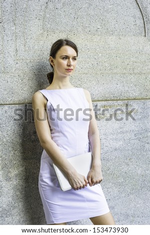 Dressing in a white sleeveless work clothes, holding a small computer,  a businesswoman is standing outside, waiting and thinking / Waiting Outside