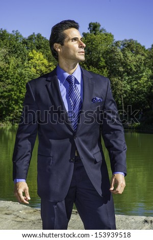 Dressing in a blue suit, blue shirt and blue pattern tie, a middle age businessman is standing by a lake and looking forward. / Looking forward