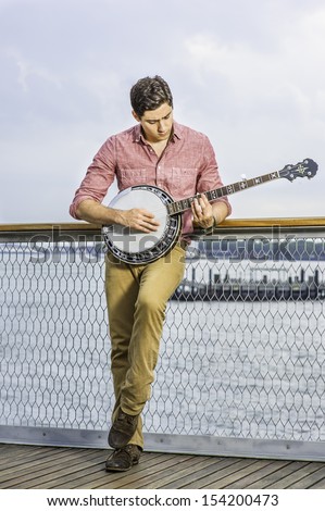 Dressing in light red shirt, dark yellow trousers and brown shoes, hanging his head and stepping his foot, a young musician is playing a banjo on the deck / Play Music