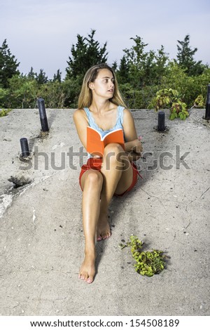A pretty blonde girl, dressing in light blue tank top, red shorts and barefoot, is reading outside on a old construction site. / Reading Outside