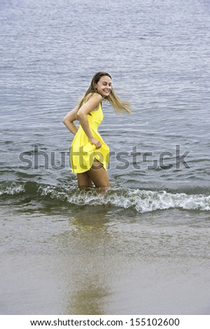 Dressing in a bright yellow dress,  a young pretty girl is wading in water on the beach, smilingly looking back / Happy Girl Wading in Beach