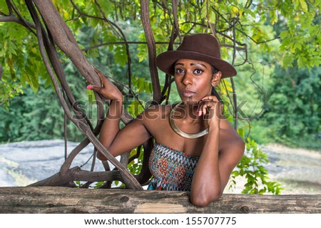 Wearing a brown outback hat,a golden neck ring, and one arm supporting her head, a pretty black girl is standing outside in a small woods  / Portrait of  Pretty Black Girl