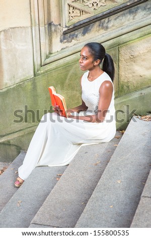 Dressing in a long white dress, sitting on steps,  a young pretty black girl is reading a red book outside. / Reading Outside