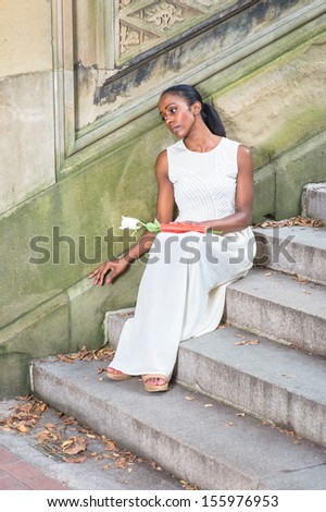 Holding a white rose and a red book on her leg, sitting on steps and leaning on the wall,  a young pretty black girl is relaxing outside and thinking. / Thinking Outside