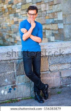 Dressing in a blue long sleeves with roll-tab Henley shirt, black jeans, leather sneakers, glasses and a wristwatch, crossing arms and one hand touching his chin,  a young guy is relaxing outside.