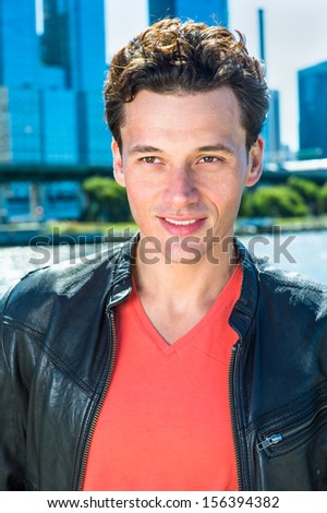 Dressing in a black leather jacket, a light red T shirt, a young handsome guy is standing by the river, smilingly looking away / Portrait of Young Guy