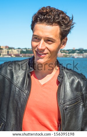 Dressing in a black leather jacket, a light red T shirt, a young handsome guy is standing by the river, smilingly looking forward / Portrait of Young Guy