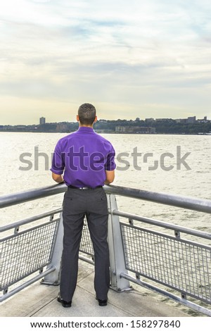 Dressing in a purple shirt, gray pants and leather shoes, a young guy is standing by a river and looking faraway.
