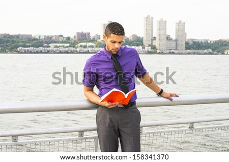 Dressing in a purple shirt, gray pants and a black tie, a young handsome college student with a little beard and mustache is standing by a river, seriouslly reading a red book. / Reading Outside