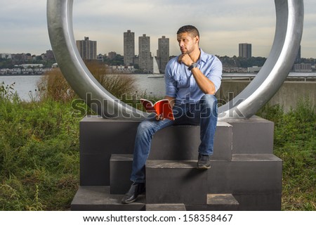 Dressing in a light blue shirt and blue jeans, one hand touching his chin, a young guy with a little beard and mustache is sitting by a river, reading a red book and thinking. / Reading Outside