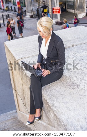 Dressing in a black suit, a young pretty blonde businesswoman is working on a computer outside / Working Outside