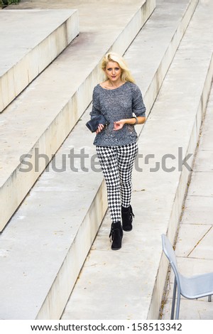 Dressing in a gray fashionable sweater, black and white pattern pants, black boots, holding a black leather purse, a young blonde girl is walking on steps. / City Girl