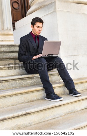 Dressing in a red undershirt, a black blazer, black jeans and a black tie, a young handsome businessman is working on a computer outside an office. / Working Outside
