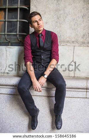 Dressing in a red shirt, a black vest, black jeans and a black tie, a young handsome businessman is sitting by an old fashion style wall, relaxing. / Relaxing Outside