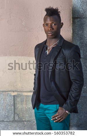 Dressing in a black blazer, green pants, a young black guy with mohawk hair is standing against the wall, confidently looking at you. / Portrait of Young Black Guy