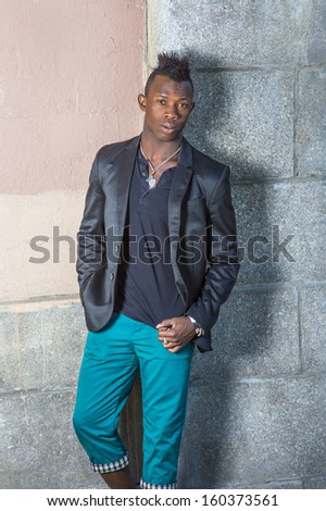 Dressing in a black blazer, a black shirt and green pants, a young black guy with mohawk hair is standing against the wall, confidently looking at you. / Portrait of Young Black Guy
