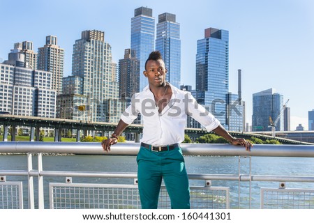 Dressing in a white shirt and green pants,  a young black guy with mohawk haircut is standing in the front of modern high buildings and looking forward. / Traveler