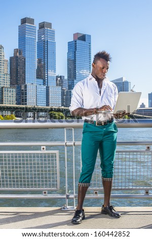 Dressing in a white shirt,  green pants and leather shoes,  a young black guy with mohawk hair is standing in the front of high buildings, working on a computer. / Leisure Time