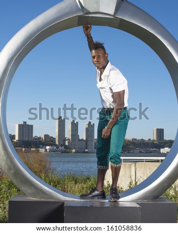 Dressing in a white shirt,  green pants and black leather shoes,  stretching one arm, a young black guy with mohawk hair is standing by a ring structure, cheerfully looking at you. / Picture in Frame