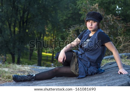 Dressing in a blue sleeveless long blouse, black leggings and boots, wearing a black corduroy cop, a young Chinese girl is sitting on rocks, seriously looking at you.