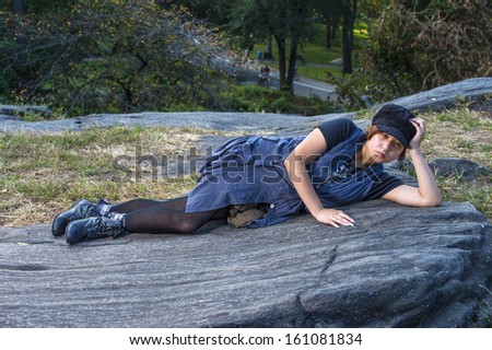 Dressing in a blue sleeveless long blouse, black leggings and boots, wearing a black corduroy cop, a young Chinese girl is lying on rocks, one hand supporting her head, into deeply thinking.