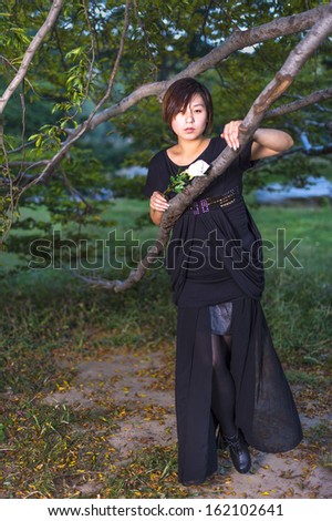A Chinese girl, dressing in dark and holding a white rose, is relying on a branch of a tree, tirelessly waiting for you. / Lonely Chinese Girl