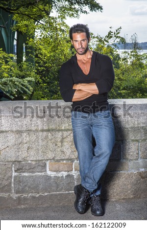 Dressing in a black sweater, blue jeans and leather shoes, crossing arms, a handsome, sexy, middle age guy with mustache and beard is leaning against the half wall fence, seriously looking at you.