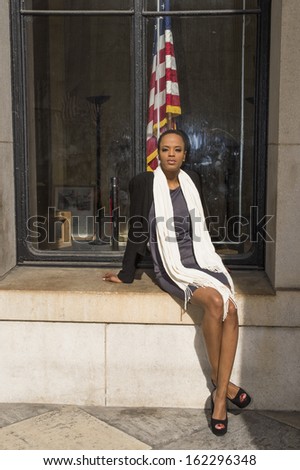 Dressing in a black jacket,  a gray dress, open toes shoes and a long white scarf, a young black businesswoman is sitting on the window with a flag in the background, relaxing outside.