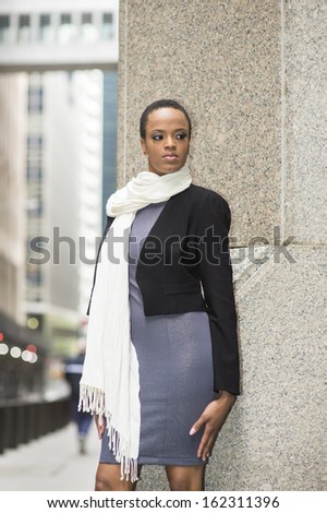 Dressing in a black woolen cropped jacket,  a gray fitted dress, and a long white scarf,  a young black businesswoman is standing outside the office building and waiting for you. / Greeting Outside